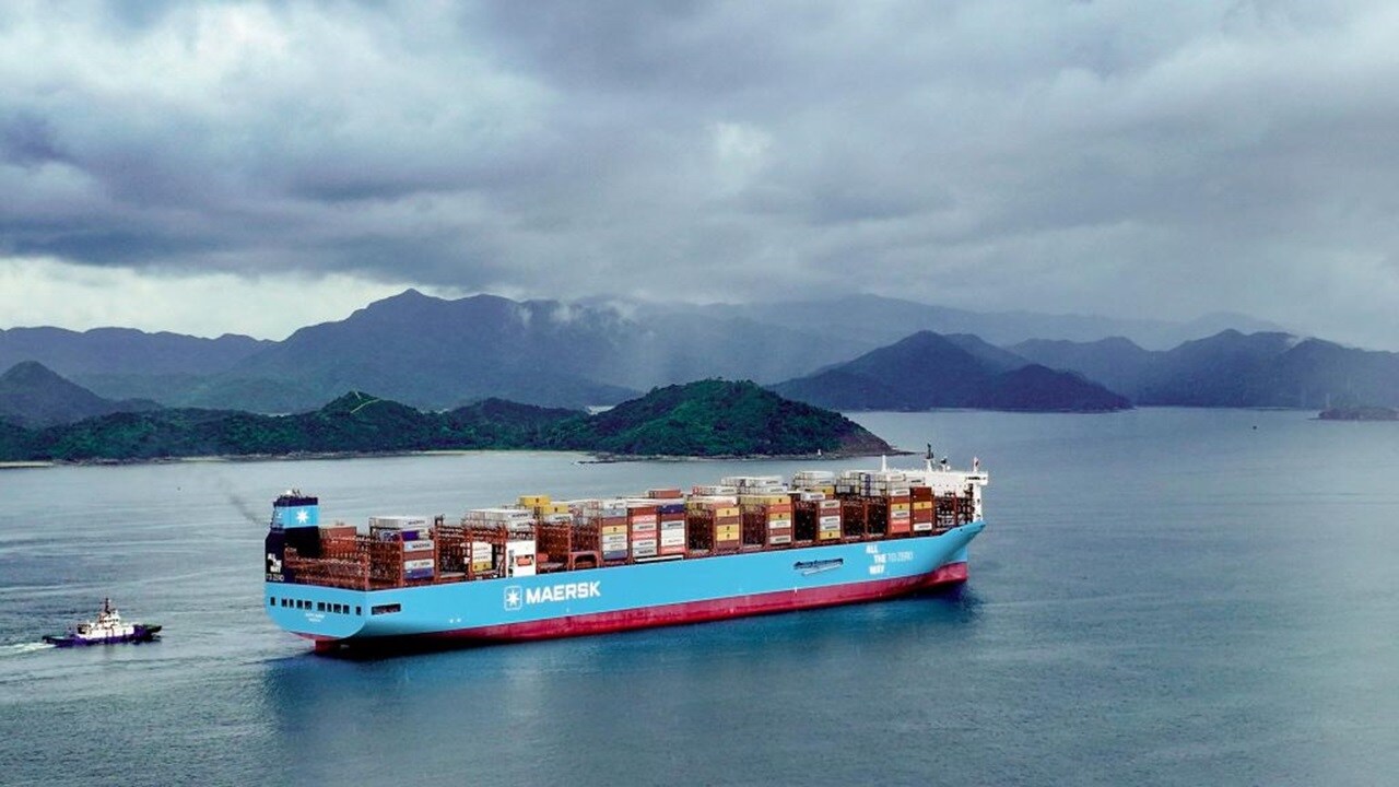 Maersk container ship carrying stacked cargo containers sailing on calm water near a mountainous coastline under cloudy skies.