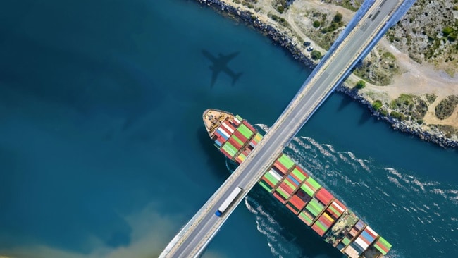 A cargo ship passes under a bridge as an airplane’s shadow reflects on the water, symbolizing global trade and multimodal logistics.