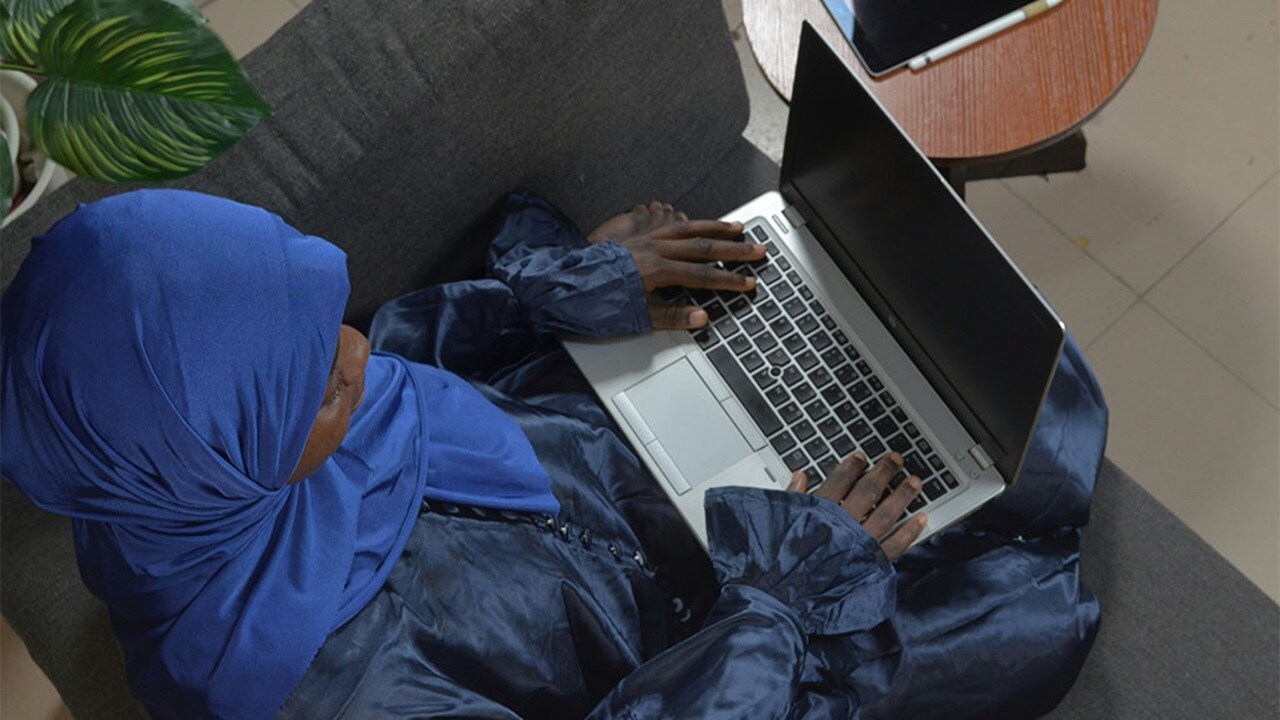 A woman in a blue robe sits on a couch, using a laptop and looking focused on her work.