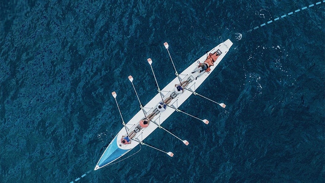 Aerial view of a team rowing boat moving across open water with multiple oars in motion.