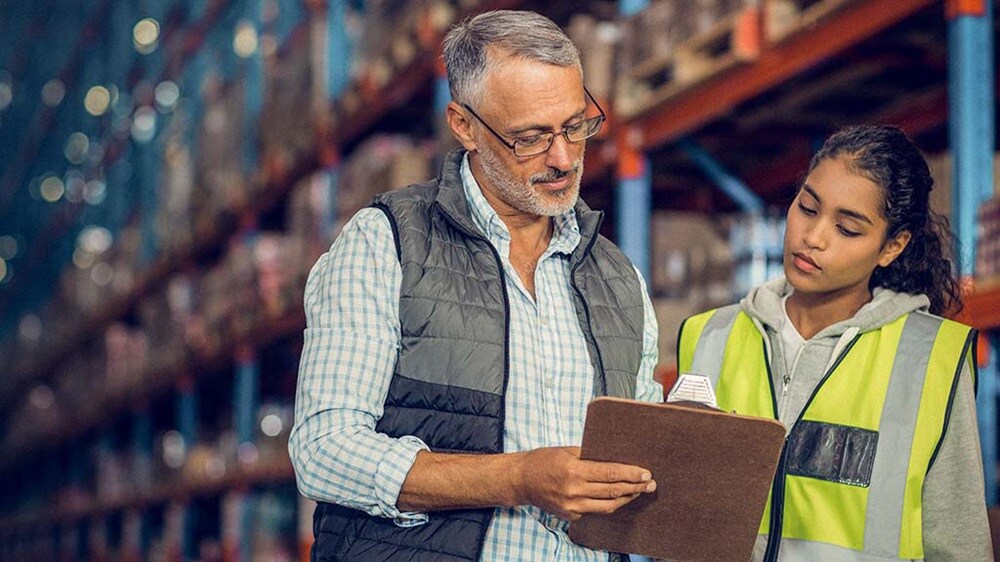 A man and woman stand in a warehouse, reviewing a clipboard together, surrounded by shelves and boxes.