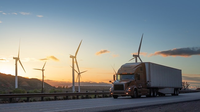 A semi-truck drives past wind turbines at sunset, symbolizing sustainable logistics and renewable energy.