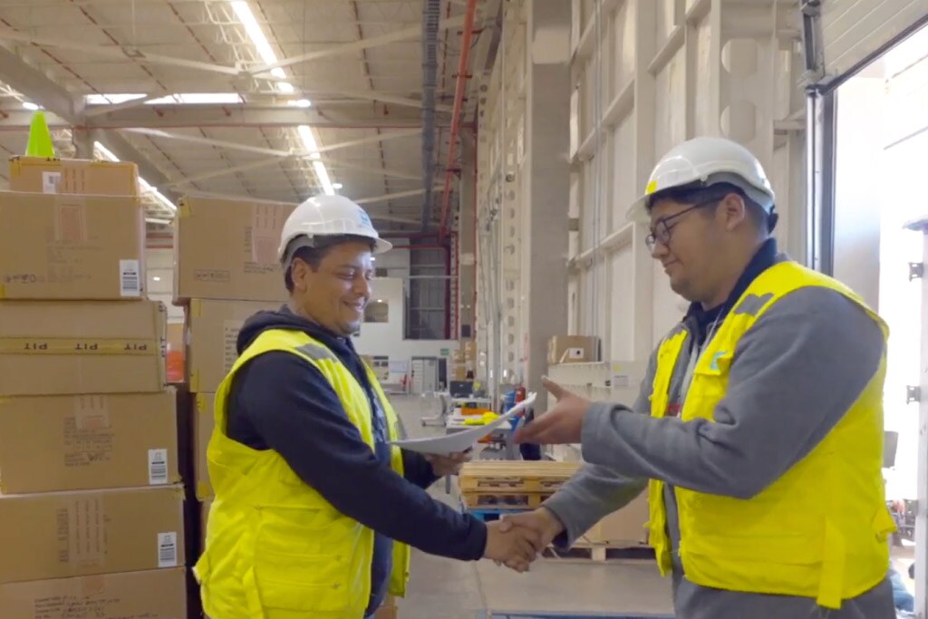 Two warehouse workers in safety vests and helmets shake hands while exchanging documents.