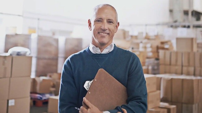 A smiling man standing with a writing board against the background of a warehouse with cardboard boxes.