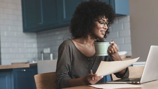 A smiling woman holding a coffee mug in one hand and a paper in another, sitting at her kitchen desk and looking at the laptop.