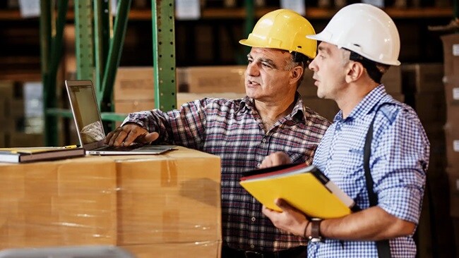 Two men in yellow safety hats viewing a laptop in a warehouse.