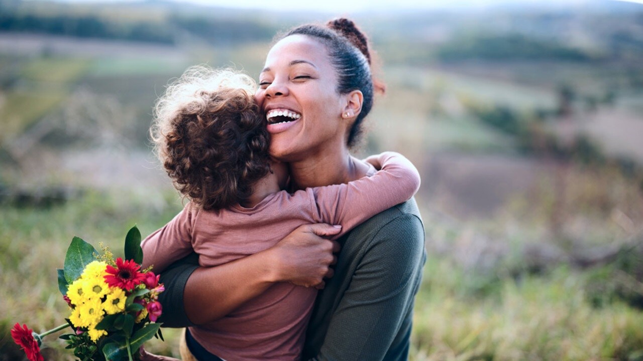A woman and her child embrace warmly in a sunlit field, surrounded by tall grass and wildflowers.