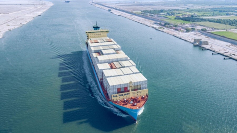 A large container ship navigates down a river, surrounded by water and greenery along the banks.