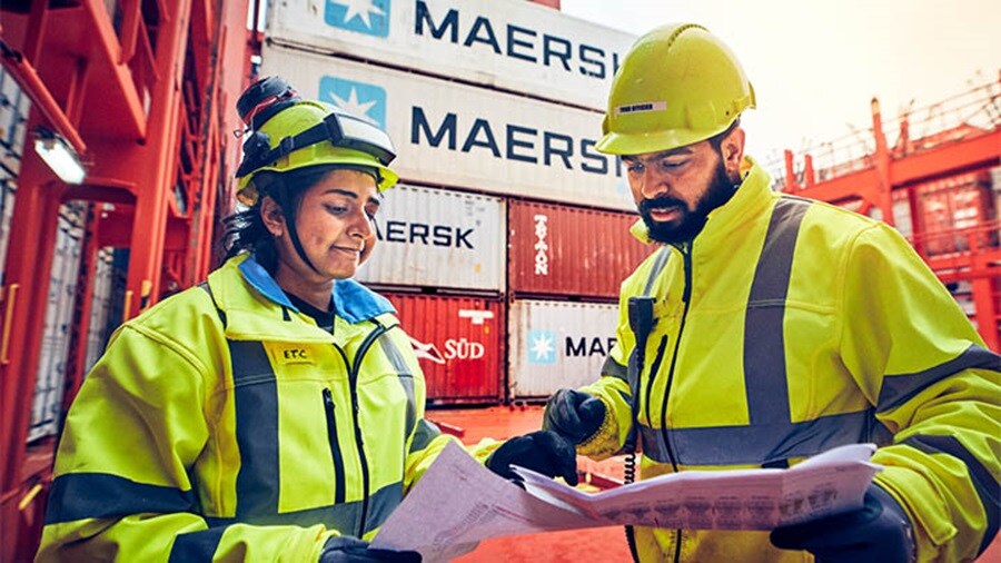 Two Maersk workers in safety gear reviewing documents at a busy shipping container yard.