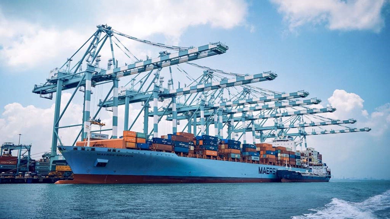 A large container ship navigates the water, surrounded by towering cranes at a busy port.