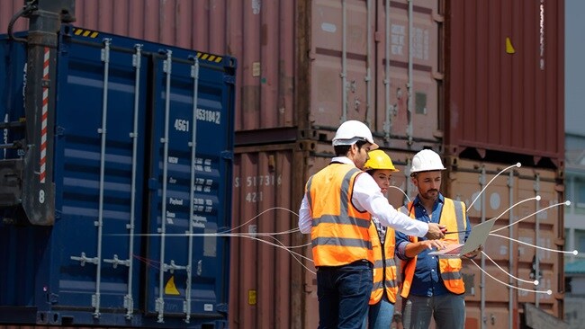 Three men in orange vests stand beside colorful shipping containers at a logistics yard.