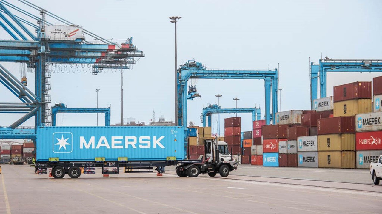 A truck parked in front of a busy container terminal, with shipping containers stacked in the background.