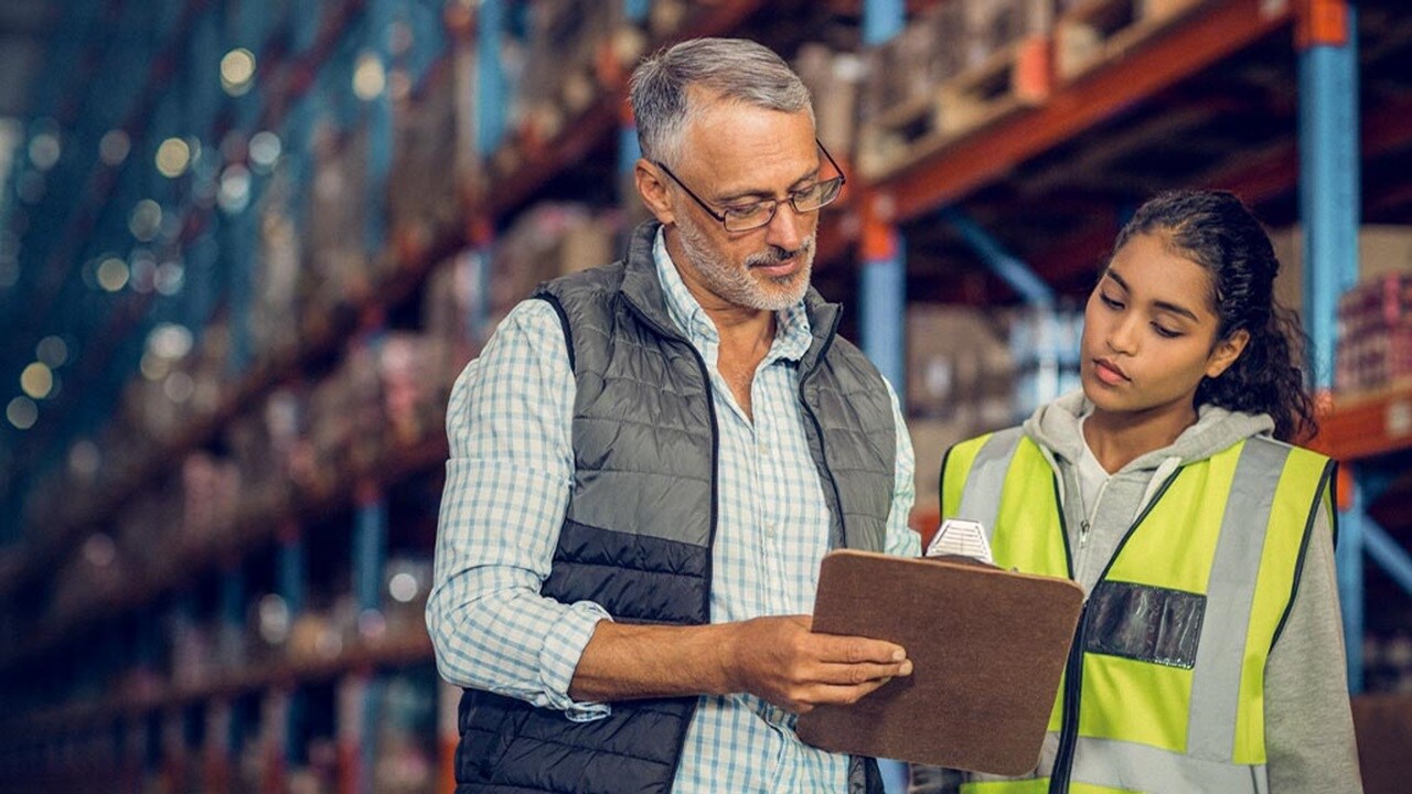 A man and woman stand in a warehouse, holding a clipboard and discussing inventory or logistics.