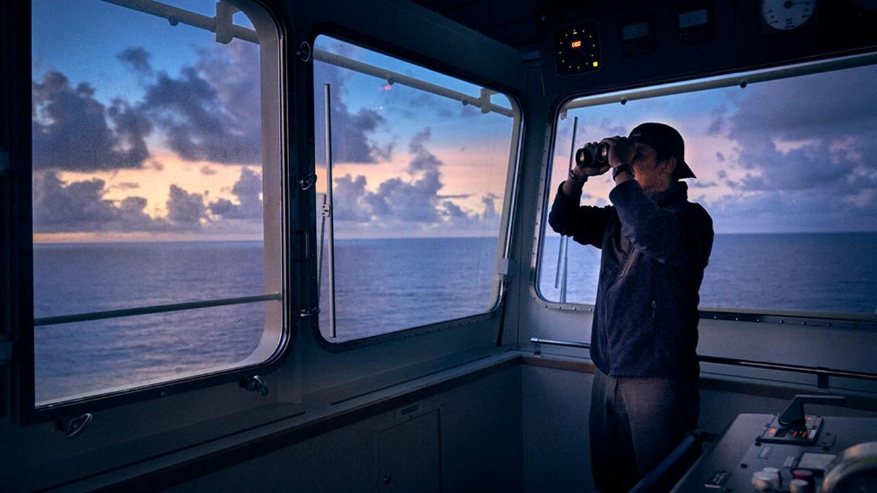 A man gazes out the window of a ship, observing the vast ocean and distant horizon.