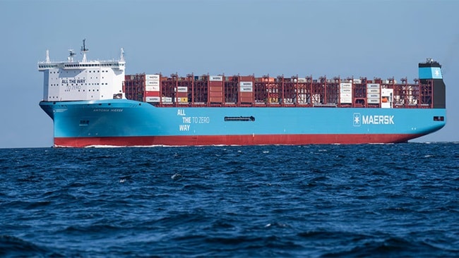 A large Maersk container ship, painted blue and red and loaded with shipping containers, sails on the open sea under clear skies.