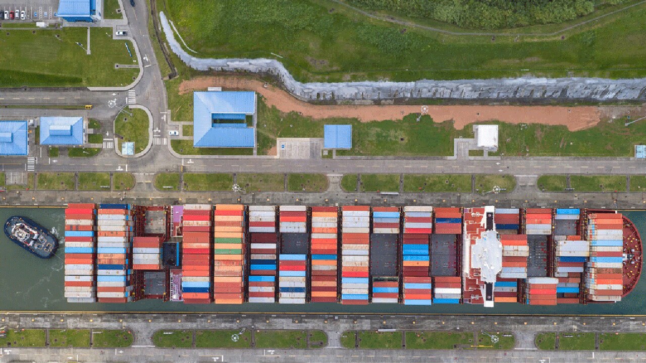 Overhead perspective of a container ship traveling in a canal, showcasing its cargo and the surrounding landscape.