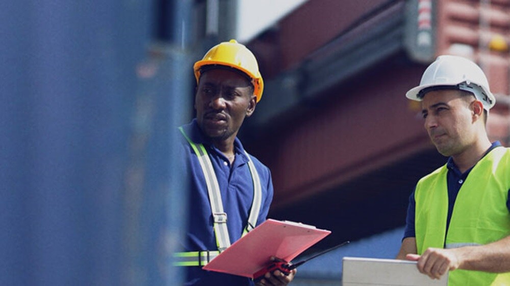 A couple of male workers in protective gear looking at a cargo container at a terminal.