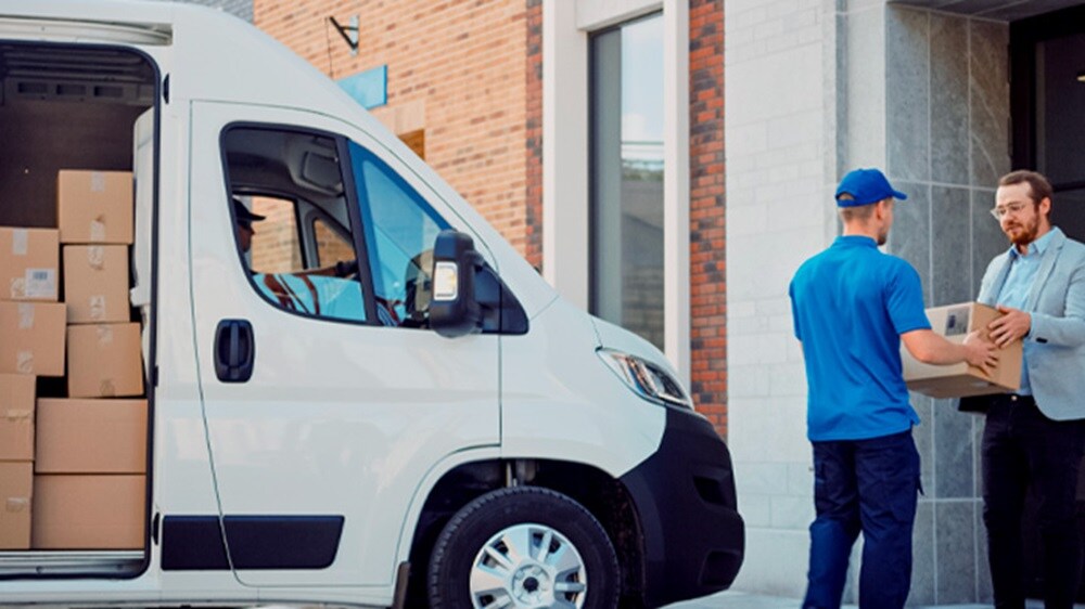 E-commerce last-mile delivery – A delivery man handing a package to a customer with a truck containing packages behind him.