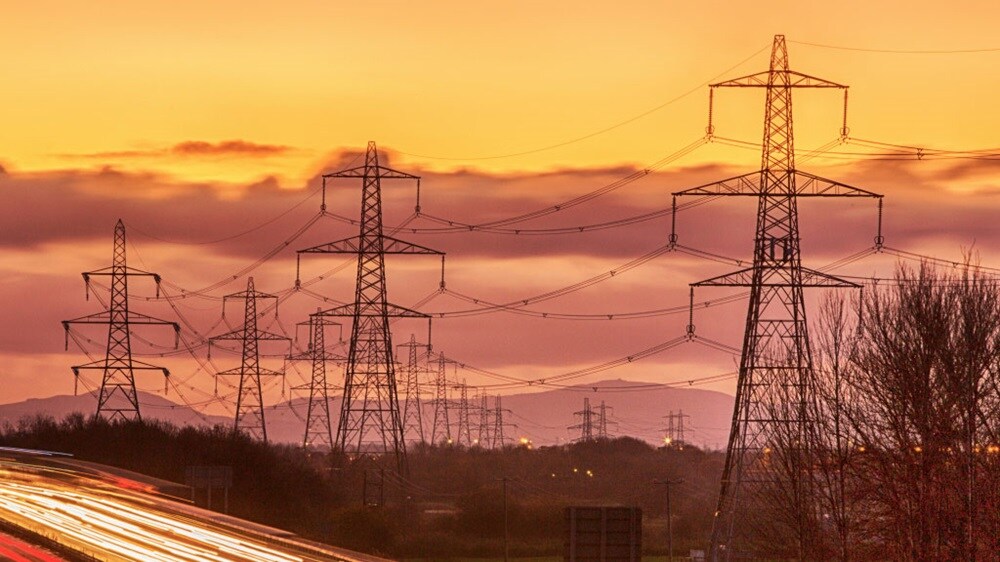 Silhouetted power lines against a vibrant orange sunset, with car light trails in the foreground and trees on the edge.