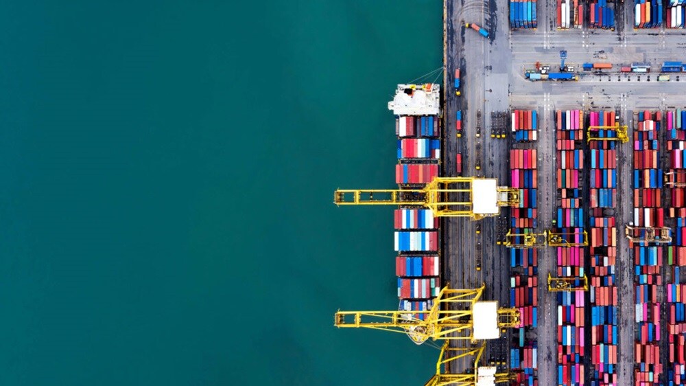 Vista aérea de un puerto de orígenes con grúas cargando contenedores de transporte y pilas organizadas a lo largo de la terminal