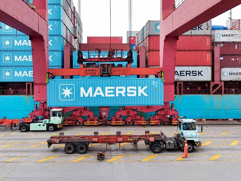 Maersk shipping container being lifted by a gantry crane at a busy port terminal with trucks below