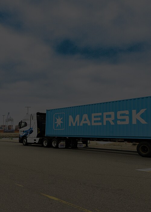 A blue Maersk truck is parked on a road near an industrial port, with shipping containers and cranes visible in the background under a cloudy sky.