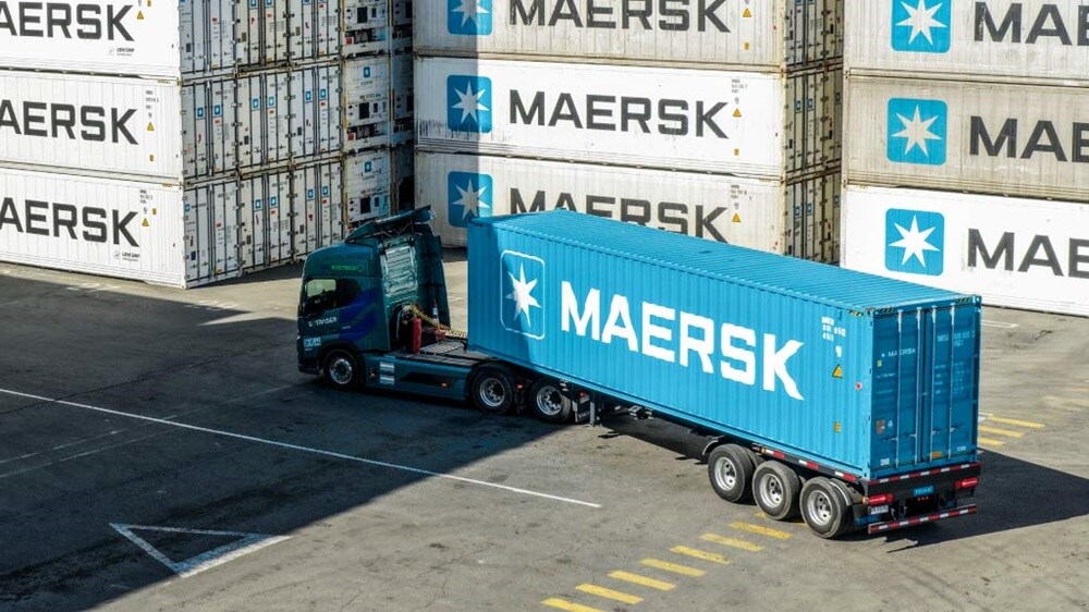 A container truck carrying a shipping container through a logistics terminal surrounded by stacked refrigerated and dry cargo containers