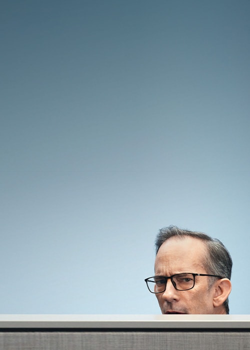 A man wearing glasses looks out from behind a cubicle wall, representing curiosity and teamwork in a business setting